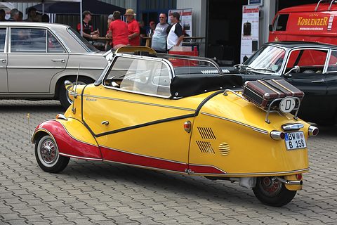 A yellow Messerschmitt KR200 Cabrio-Limousine with red lower panels, its fabric roof folded and a suitcase on the rear rack, parked on cobbles at a show. Opens a larger view
