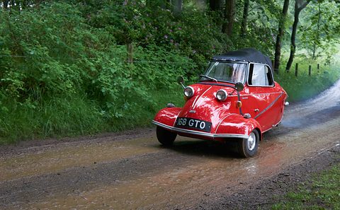 Red three-wheeler with black soft top driving through puddles on a muddy track between trees. Opens a larger view