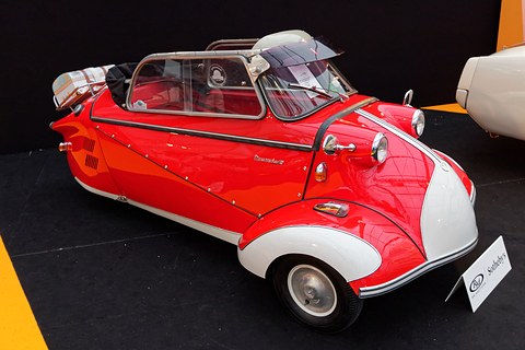 Red three-wheeler with white front wings, soft top folded, on a black auction stand. Opens a larger view