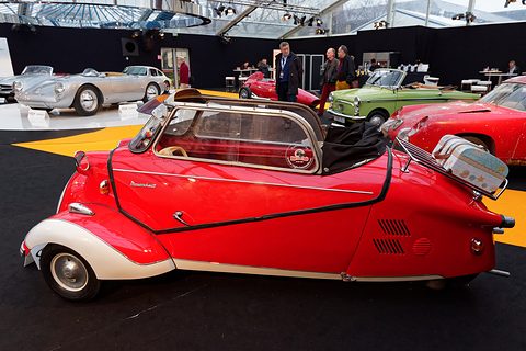 Red three-wheeler with white wheel arches side-on, small cases strapped to the rear rack. Opens a larger view