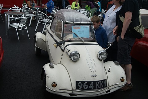 White bubble car, registration 964 KKN, surrounded by browsing visitors and cafe chairs. Opens a larger view