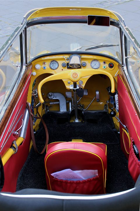 Yellow dashboard and handlebar above black floor mats and pedals, a red seat behind. Opens a larger view
