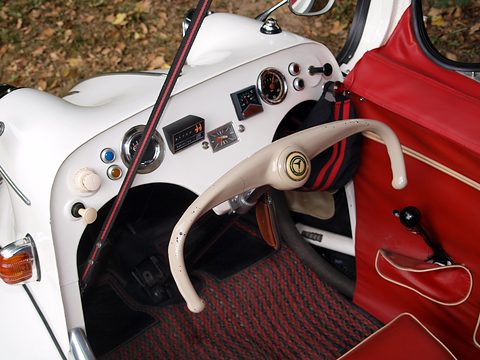 Close view of a white dashboard with dials and switches, cream handlebar and red door trim. Opens a larger view