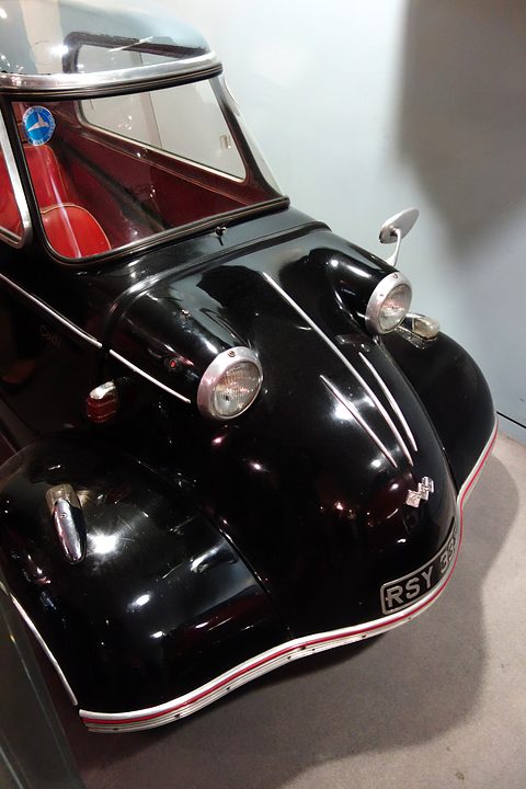 Black bubble car with chrome trim and headlamps seen head-on in a white gallery. Opens a larger view