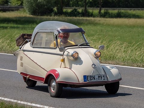 Cream bubble car with red trim and luggage on its rack on a road beside a meadow, driver visible. Opens a larger view