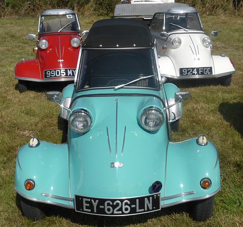 Head-on view of a turquoise four-wheeled bubble car with a black fabric top, a red and a white three-wheeled bubble car behind. Opens a larger view