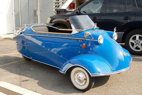 Bright blue open three-wheeler with whitewall tyres and a chrome rack in a car park. Opens a larger view