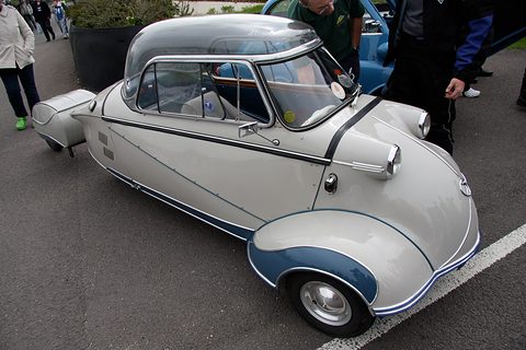Grey bubble car with blue wheel arches on tarmac, people standing behind. Opens a larger view