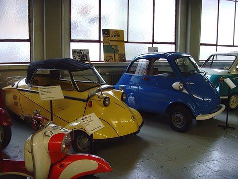 Yellow three-wheeler with black soft top next to a blue Isetta by tall windows. Opens a larger view