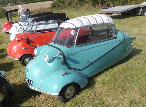 Turquoise three-wheeler with a striped fabric top in front of a red and a white Messerschmitt. Opens a larger view