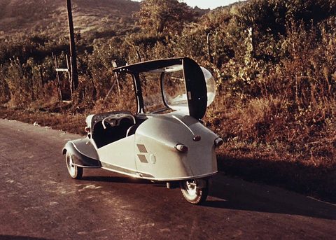 A faded colour photograph of a pale Messerschmitt parked at a roadside with its canopy swung open. Opens a larger view