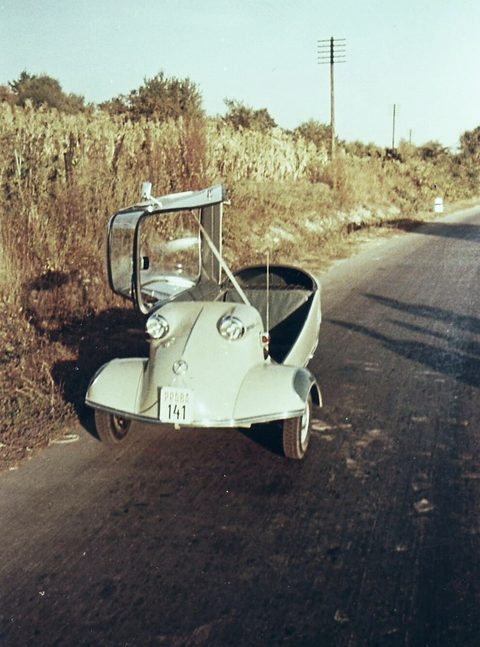 A faded colour photograph of a pale Messerschmitt seen head-on on an empty country road, canopy open. Opens a larger view