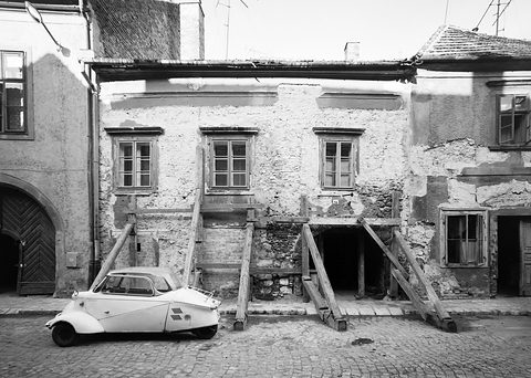 Black-and-white photograph of a small pale Kabinenroller parked in front of a shabby two-storey house propped up with timber struts