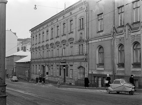 A wintry city street of tall plastered buildings, pedestrians in coats and a small bubble-canopied three-wheeler at the kerb. Opens a larger view