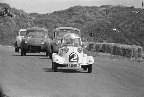 Black-and-white photograph of a small four-wheeled bubble car numbered 2 leading two saloons round a circuit bend below dunes.