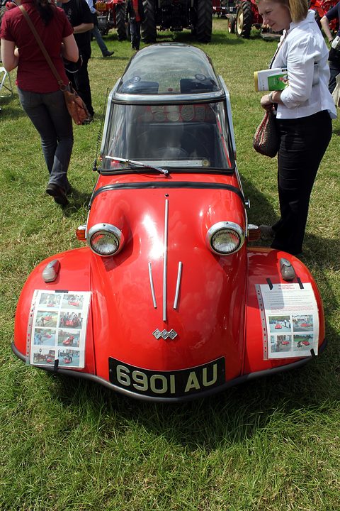 Red bubble car head-on on grass, papers taped to both front wings, visitors around. Opens a larger view
