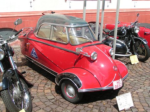 Red bubble car on cobbles between black BMW motorcycles, a placard on the ground. Opens a larger view