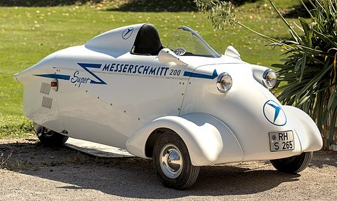 A white streamlined single-seat three-wheeler with a headrest fairing, blue lettering and a small screen, on gravel. Opens a larger view