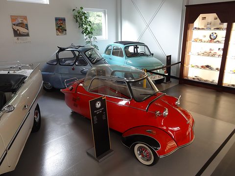 Red bubble car with chrome hubcaps beside a black sign, other microcars behind. Opens a larger view