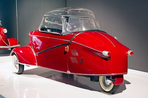 Red tapering tail with twin lamps and louvres on a white plinth against a grey wall. Opens a larger view