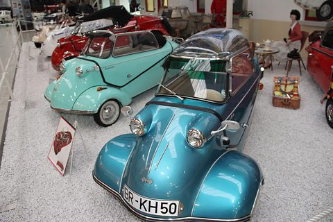Teal bubble car, plate GR-KH 50, facing the camera, a turquoise roadster and picnic basket behind. Opens a larger view