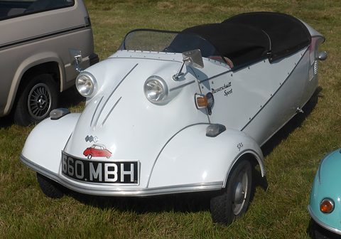 A white three-wheeler with no canopy, a low curved screen and black tonneau, parked on grass. Opens a larger view