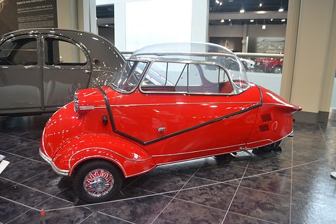 Red tandem bubble car side-on on dark tiles, a grey car behind. Opens a larger view