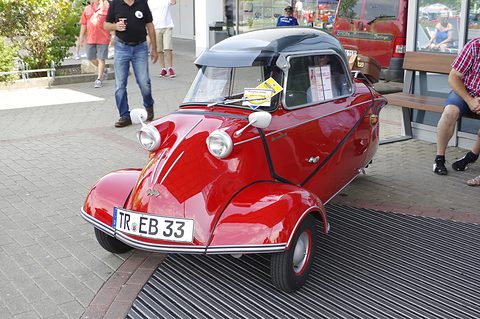 Red bubble car with dark canopy on a doormat-like grid, visitors around it. Opens a larger view
