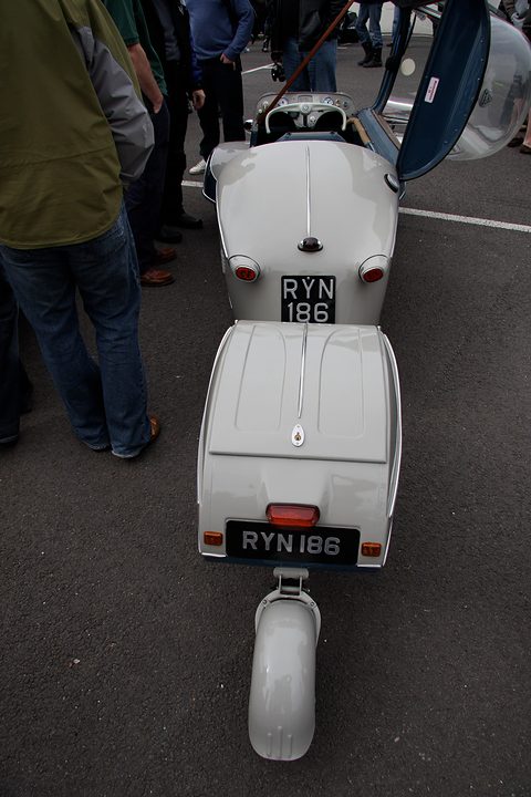 Grey bubble car and a small grey trailer from behind, plate RYN 186, visitors’ legs either side. Opens a larger view