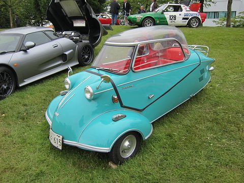 Turquoise bubble car with red interior on grass beside a silver sports car. Opens a larger view