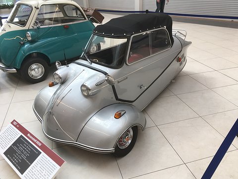 Silver three-wheeler with black soft top on white tiles beside a teal Isetta, a display board in front. Opens a larger view