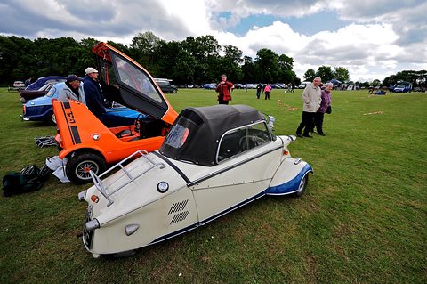 White three-wheeler with blue trim and black soft top beside an orange car with its canopy raised. Opens a larger view