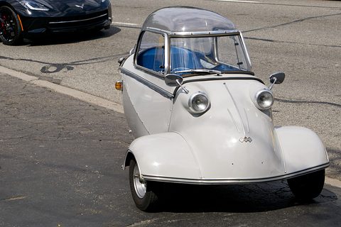 White bubble car with blue seats and FMR badge on cracked tarmac, a black Corvette behind. Opens a larger view