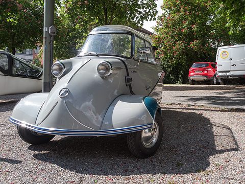 Grey bubble car with blue-green wings facing the camera on gravel beside a pole. Opens a larger view