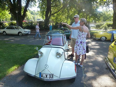 White bubble car head-on in a park, a couple standing beside it. Opens a larger view