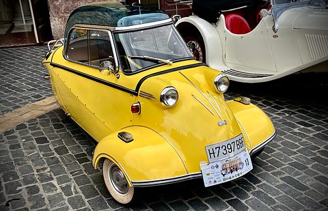 Yellow bubble car with dark-tinted canopy parked on grey cobbles beside a white roadster. Opens a larger view