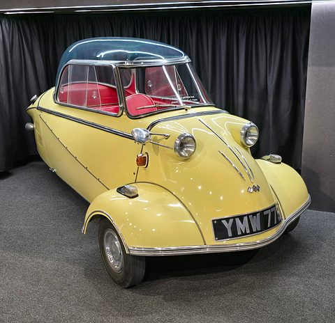 Yellow bubble car with red interior on a dark display stand against a black curtain. Opens a larger view