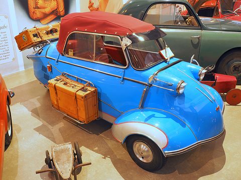 Blue three-wheeler with red soft top, suitcases on its rack and beside it, a trailer behind. Opens a larger view