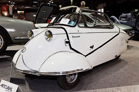 White open three-wheeler with black trim line, canopy-style hood frame raised, in a show hall. Opens a larger view