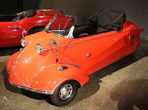 Orange-red open microcar with folded black hood and chrome trim in a dim museum. Opens a larger view