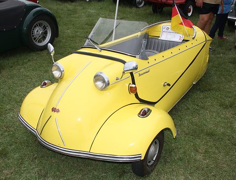 Yellow open three-wheeler with a small windscreen and a German flag, parked on grass. Opens a larger view