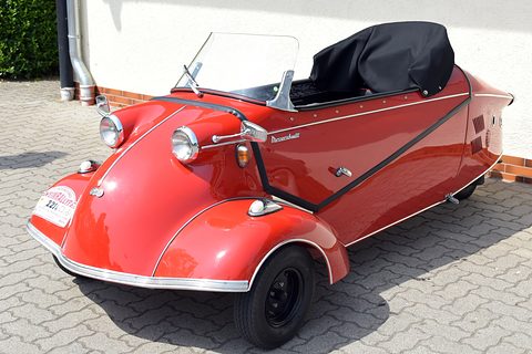 Red open three-wheeler with folded black hood and chrome trim on paving beside a white wall. Opens a larger view
