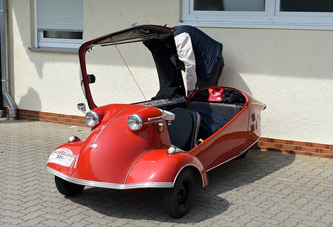 Red open microcar with a black folding hood frame lifted up and a jacket on the seat. Opens a larger view