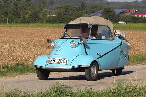 Light-blue three-wheeler with tan hood, registration M-KR 2001, on a farm road beside a field. Opens a larger view