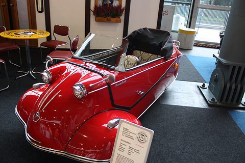 Red three-wheeler with black hood raised, an information card on a stand in front, museum interior. Opens a larger view