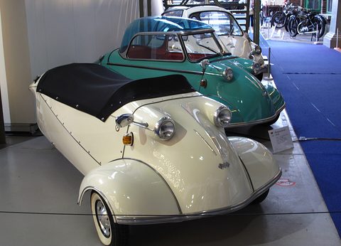 Cream open three-wheeler with a black tonneau, a turquoise Messerschmitt behind, on a blue carpet. Opens a larger view