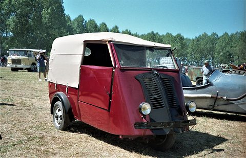 Faded maroon three-wheeled van with a cream canvas back and black louvred nose, parked on dry grass in sunshine.