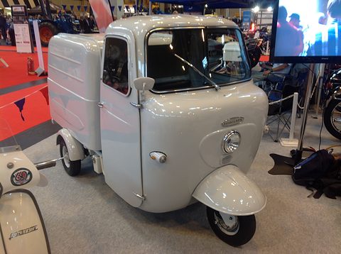 Pale grey three-wheeled van with a rounded cab and a box body on a show stand. Opens a larger view