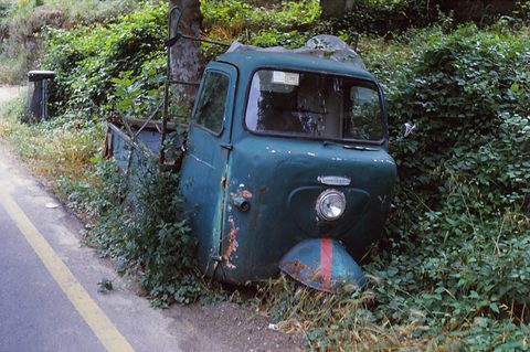 Rusting blue three-wheeled pick-up with a single front wheel half buried in ivy beside a road. Opens a larger view