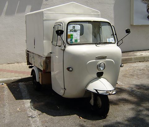 White three-wheeled cab-over van with a box body and a single front wheel parked by a wall.